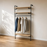 Étagère-penderie murale en tuyaux noirs, trois tablettes en bois et chemises suspendues à la barre, mur beige et parquet