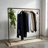 Arara de roupa industrial em tubos de canalização pretos numa loja com parede de tijolo branco, com planta pendente e lâmpada